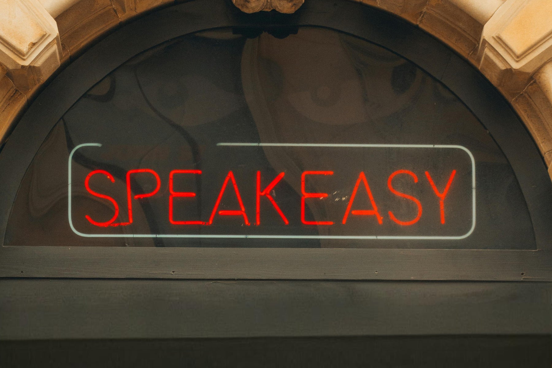 A red neon sign reading "SPEAKEASY" in a window above a doorway with ornate stonework.