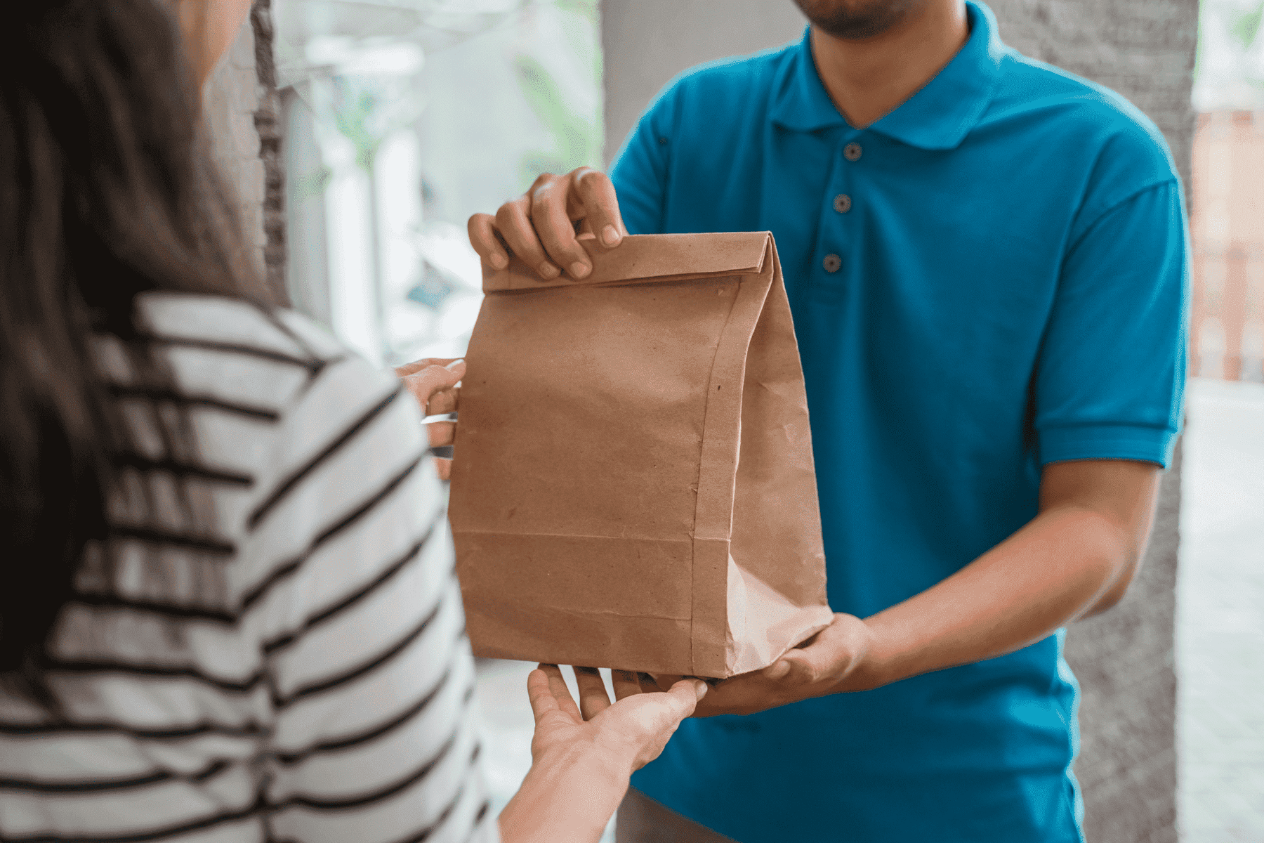 Someone receives a food order in a paper bag from a delivery driver in a blue polo t-shirt